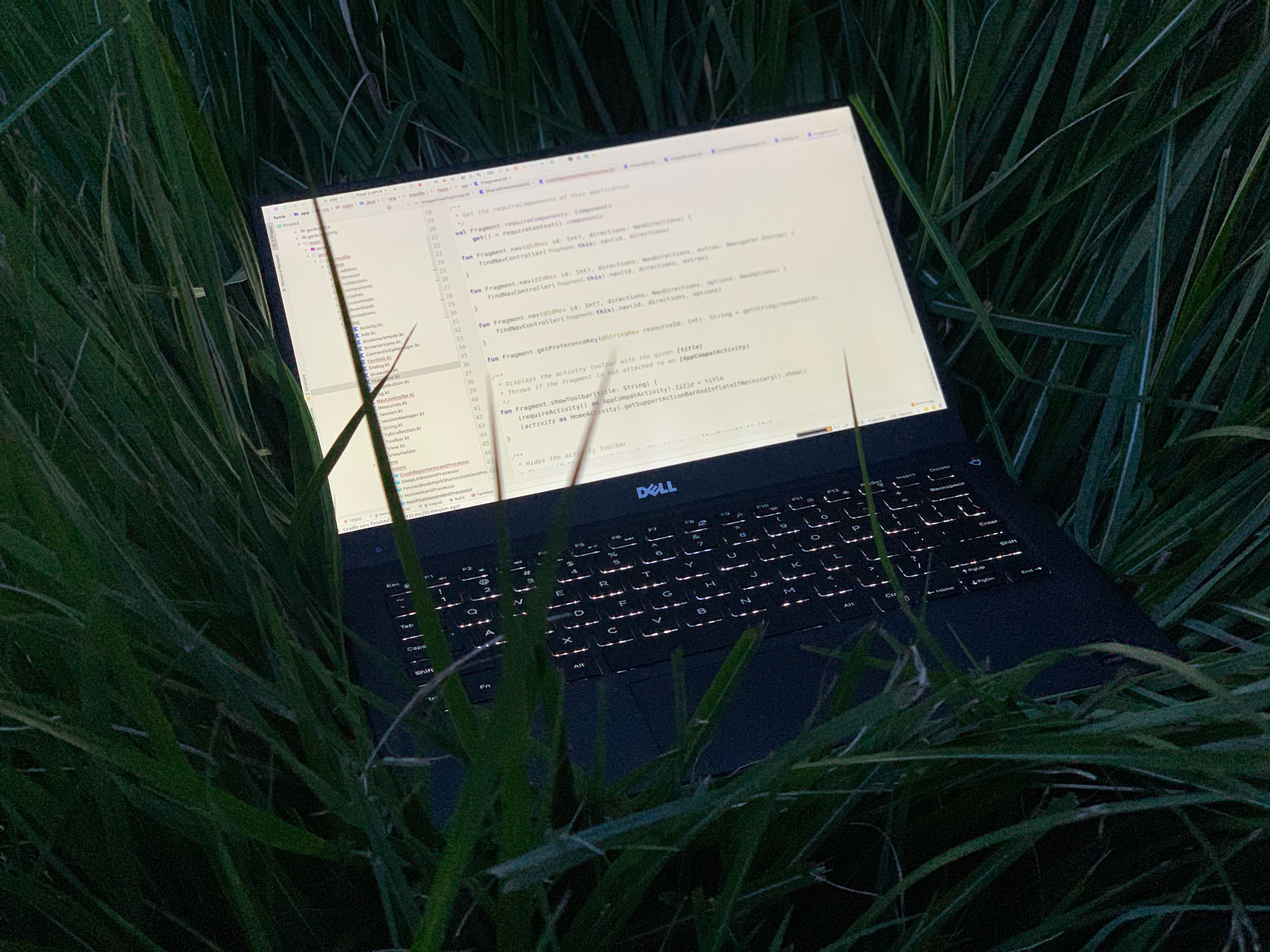 A laptop resting in some deep grass. The laptop is displaying kotlin code from the Firefox Fenix project.
