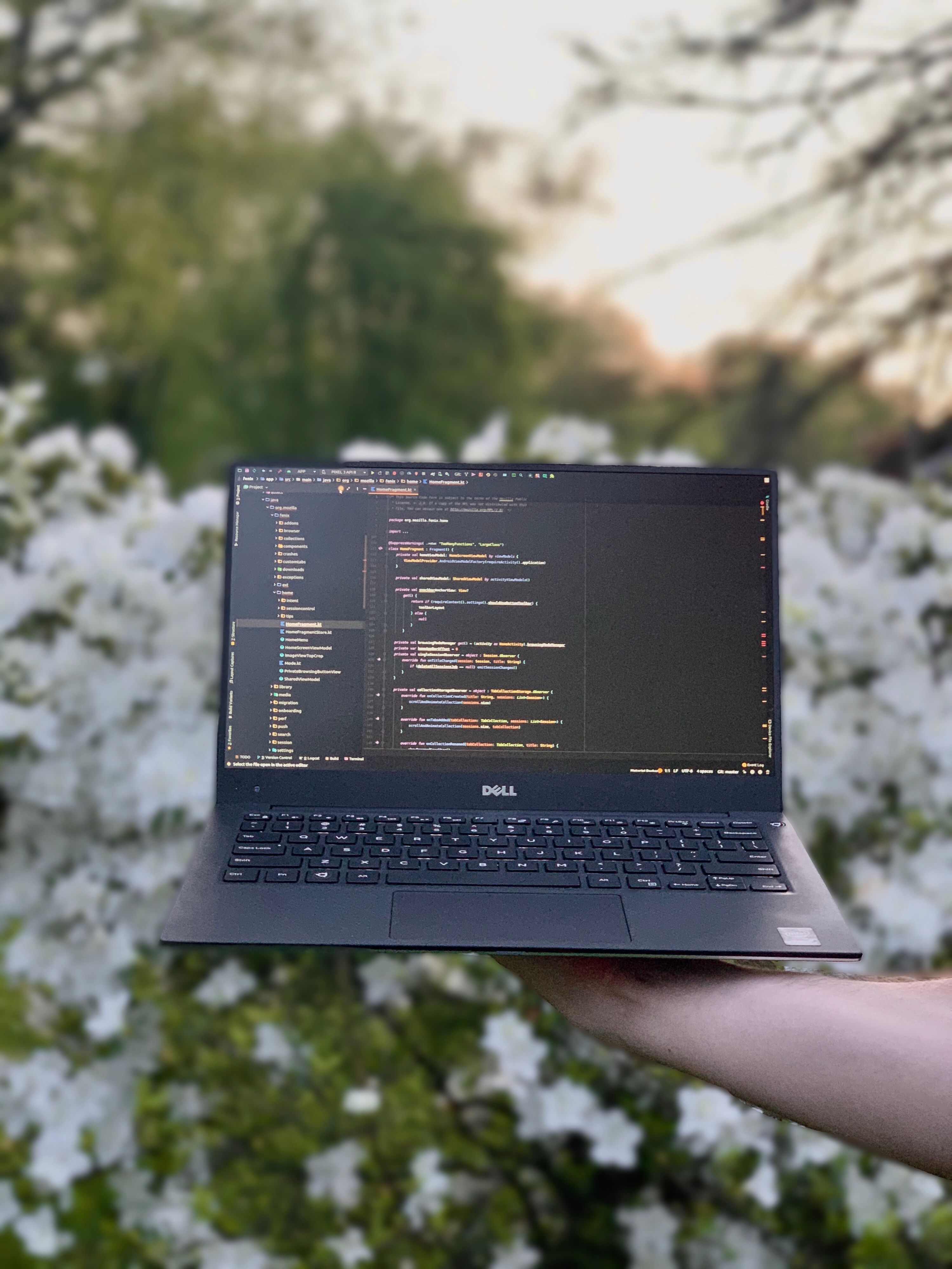 Holding up a laptop in front of white flowers. The laptop is displaying kotlin code from the Firefox Fenix peoject.