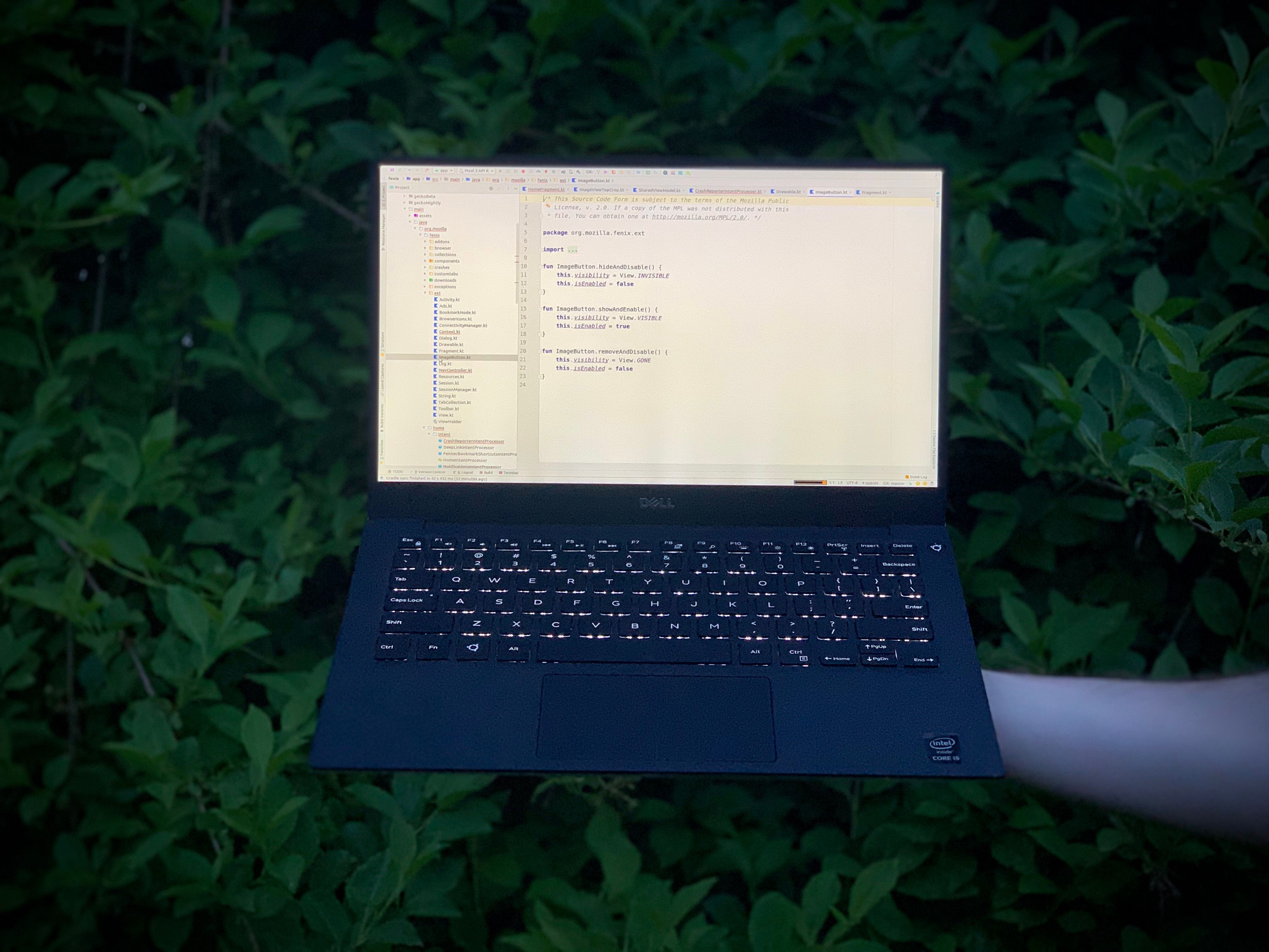 Holding up a laptop with a leafy green background. The laptop is displaying kotlin code from the Firefox Fenix project.