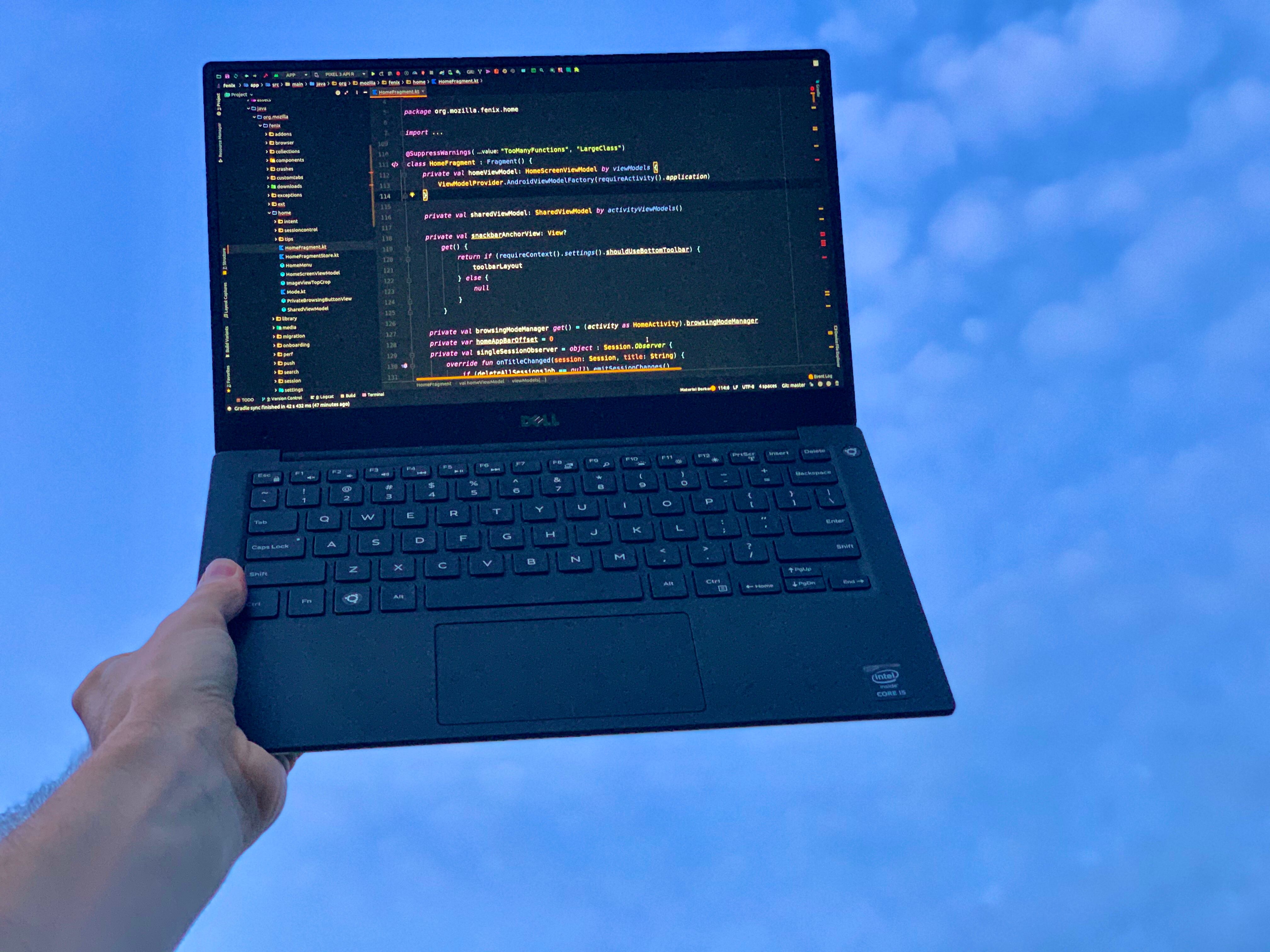 Holding up a laptop with one hand using the sky as a backdrop. The laptop is displaying kotlin code from the Firefox Fenix project.